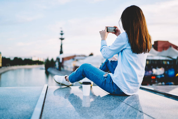 Joven mujer milenaria usando aplicación de teléfono inteligente para hacer fotos mientras descansa en el entorno urbano, vista posterior de la blogger hipster femenina en el video casual de rodaje de ropa en el teléfono móvil digital - Foto, Imagen
