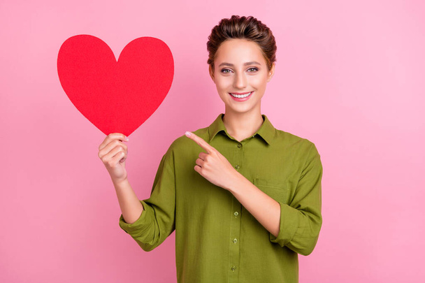 Photo of sweet shiny young woman dressed green shirt smiling pointing finger red heart empty space isolated pink color background - Foto, Imagem