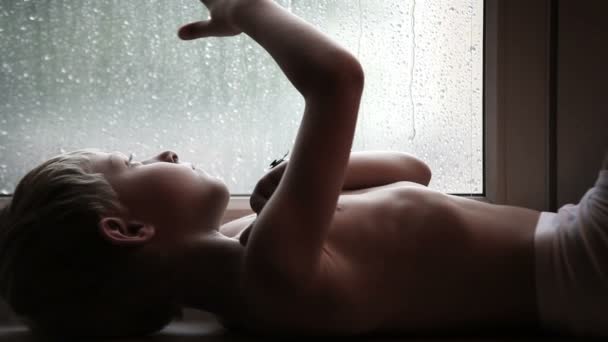 Sad boy on the windowsill watches the raindrops on the glass - Πλάνα, βίντεο