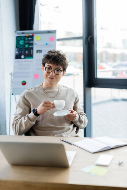 empresário em óculos segurando copo e pires perto de laptop borrado e notebook no escritório  - Foto, Imagem