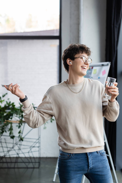 Usmívající se podnikatel ve sluchátku drží smartphone a sklenici vody v kanceláři  - Fotografie, Obrázek