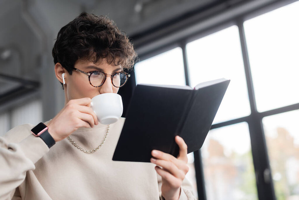 Mladý podnikatel ve sluchátku pití kávy a při pohledu na notebook v kanceláři  - Fotografie, Obrázek