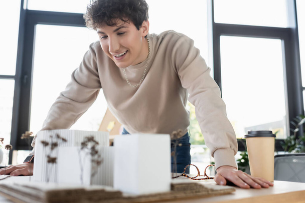 Веселый бизнесмен смотрит на модель здания возле очков и кофе, чтобы пойти в офис  - Фото, изображение