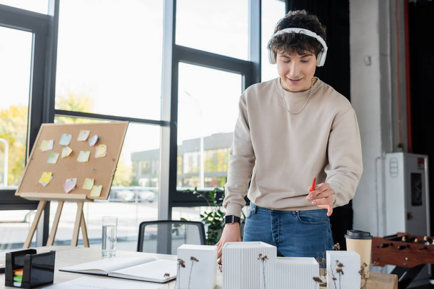 businessman in headphones holding pen near model of building and drinks in office  - Zdjęcie, obraz