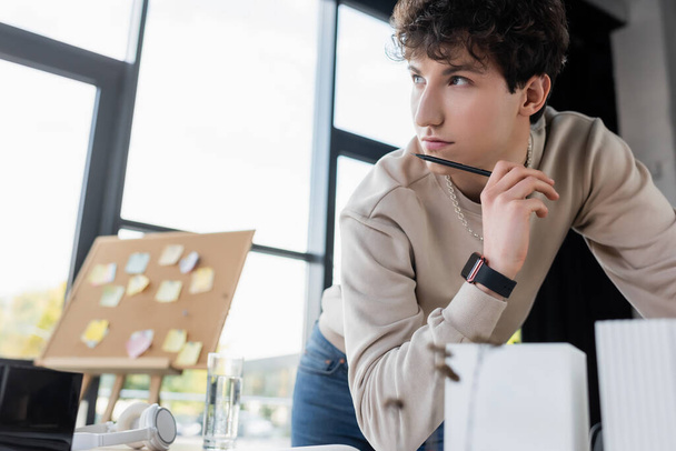 Pensive επιχειρηματίας κρατώντας στυλό κοντά θολή μοντέλο του κτιρίου και ακουστικά στο τραπέζι στο γραφείο  - Φωτογραφία, εικόνα