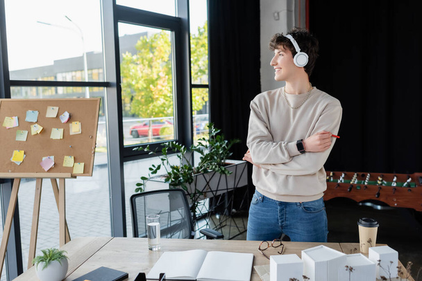 Вид збоку усміхненого бізнесмена в навушниках, що стоять біля робочого столу в офісі
  - Фото, зображення