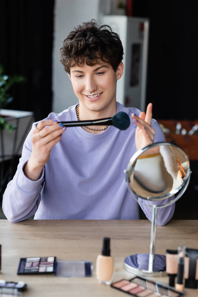 Sonriente persona transgénero sosteniendo cepillo cosmético cerca de cosméticos en la mesa  - Foto, Imagen