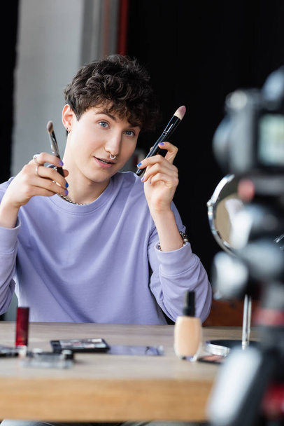 Transgender makeup artist holding cosmetic brushes near blurred digital camera  - 写真・画像