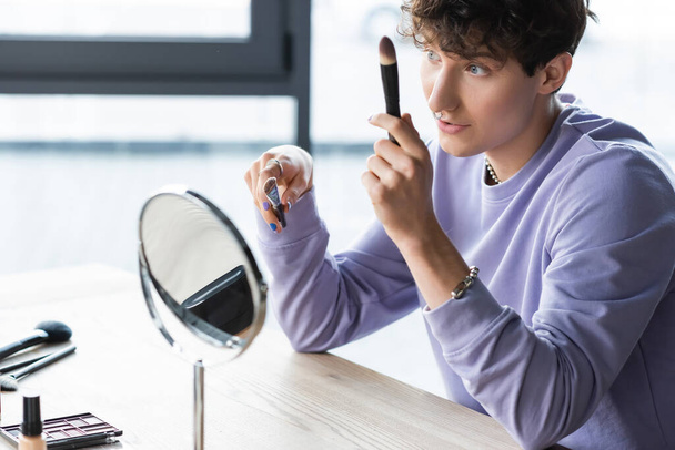 Молодой трансгендерный визажист держит косметические кисти возле зеркала  - Фото, изображение