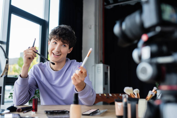 Feliz blogger transgénero sosteniendo pinceles cosméticos cerca de una cámara digital borrosa - Foto, Imagen