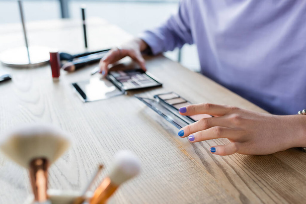 Cropped view of transgender person holding eye shadows near blurred cosmetic brushes on table  - Foto, Imagen