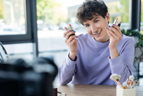 Blogger transexual positivo sosteniendo lápices labiales cerca de una cámara digital borrosa  - Foto, imagen