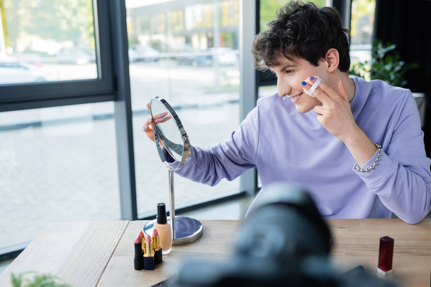 Vista laterale di una persona transgender sorridente che applica uno specchio per le orecchie in polvere per il viso e una fotocamera digitale offuscata  - Foto, immagini