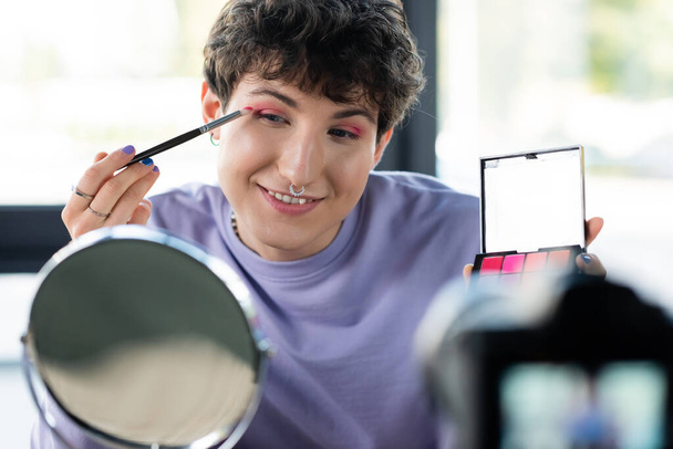 Νεαρό τρανσέξουαλ άτομο που εφαρμόζει σκιά ματιών κοντά στον καθρέφτη και θολή ψηφιακή κάμερα  - Φωτογραφία, εικόνα