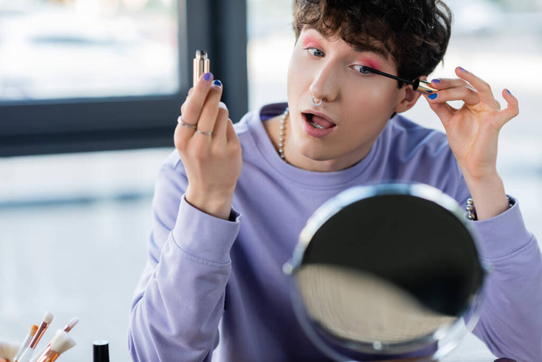 Persona transgénero aplicando rímel cerca de cepillos cosméticos y espejos  - Foto, Imagen