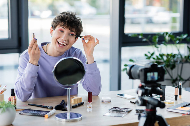 Sonriente persona transgénero aplicando rímel cerca de cosméticos decorativos y cámara digital  - Foto, imagen