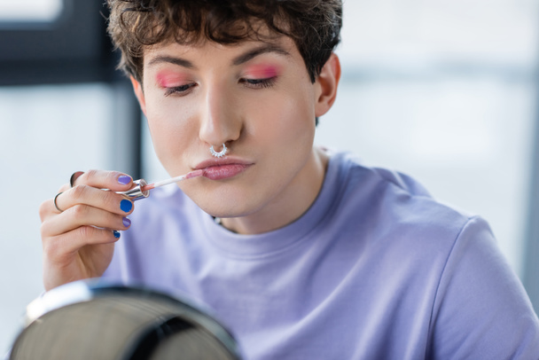 Transgênero pessoa aplicando bálsamo labial perto de espelho borrado  - Foto, Imagem