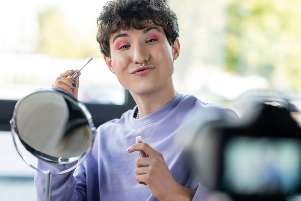 Transgenderowa osoba trzymająca balsam do ust i dąsające się usta w pobliżu lustra i aparatu cyfrowego   - Zdjęcie, obraz
