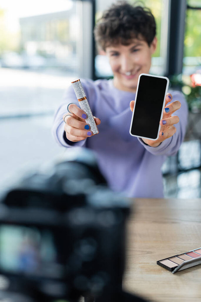 Mascara et smartphone entre les mains d'une personne transgenre floue près d'un appareil photo numérique  - Photo, image