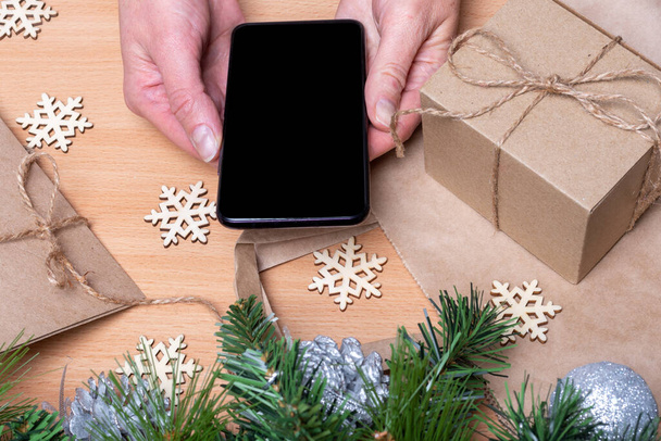 Close up hands holding mobile phone with blank black screen on wooden table with Christmas gift boxes, snowflakes, Christmas tree branches. Online shopping christmas gifts concept - Foto, Bild