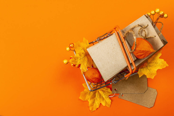 Black Friday Sale concept. Shopping basket, handmade tags, paper bags, fall maple leaves, and physalis. Orange background, trendy hard light, dark shadow, top view - Foto, imagen