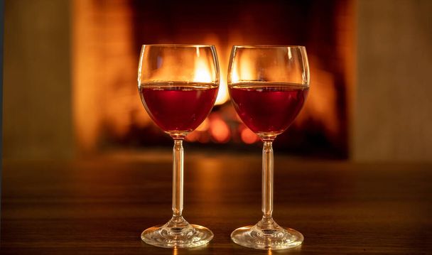 Dwie czerwone lampki wina na drewnianym stole, płonące tło kominka. Para picia, Walentynki uroczystości obiad lub koncepcja romantycznej randki - Zdjęcie, obraz