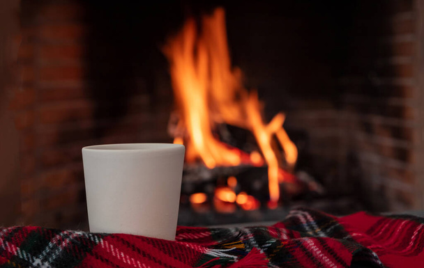 Noel tatili sıcacık evi. Kanepe üzerinde kırmızı ekose battaniyeli bir fincan kahve, yanan şömine arka planı. Köknar gevşemesi - Fotoğraf, Görsel