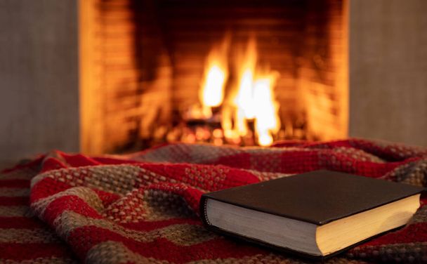 Buch auf einer Decke, Kamin brennenden Brennholz Hintergrund. Lesen und entspannen am Feuer, Weihnachtsurlaub gemütlich warme Inneneinrichtung.  - Foto, Bild