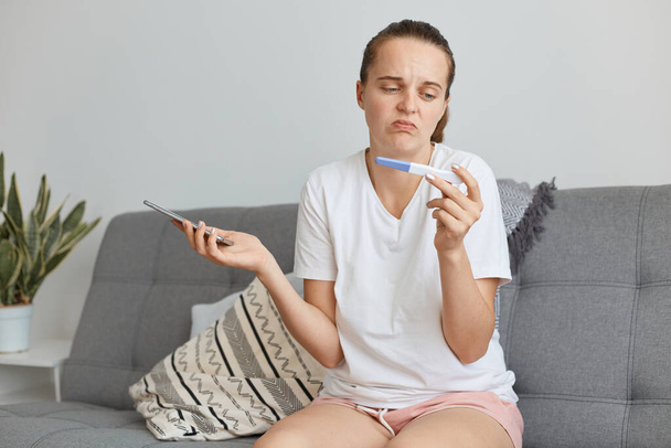 Indoor shot of attractive dark haired woman wearing white t shirt and short, sitting on sofa in living room with pregnancy test with positive result, does not know what to do. - Foto, immagini