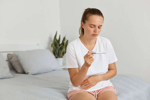 Indoor shot of sad woman with ponytail wearing casual clothing holding pregnancy test in hands, sitting on sofa in living room, having negative result, being disappointed. - Photo, Image