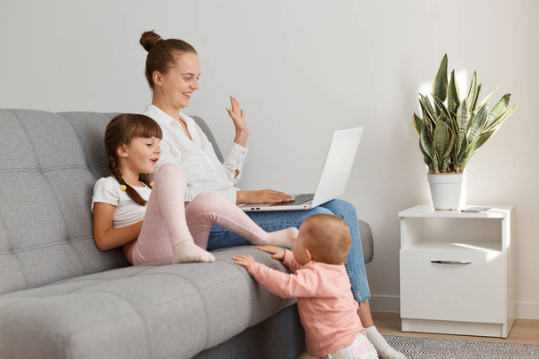 Indoor shot of optimistic smiling young woman with her little daughter with pigtails, having video call, waving hand to camera, expressing positive emotions. - Φωτογραφία, εικόνα