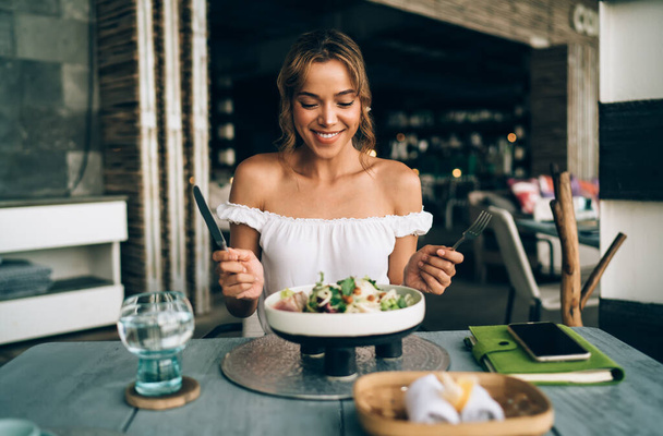 Mosolygó elegáns fiatal nő divatos felső nyitott vállak ül asztalnál hangulatos tágas kávézó evőeszközök és élvezi ízletes zöldség saláta tálalva kreatív tál - Fotó, kép