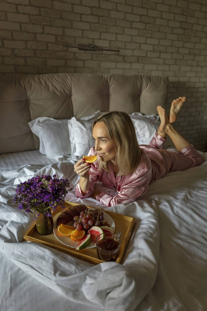 Vaalea aamiainen croissantia, hedelmiä ja kahvia tarjoillaan sängyssä kauniille nuorelle blondille varhain aamulla.. - Valokuva, kuva