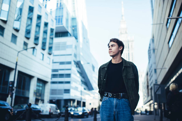Young confident male in casual outfit with hands behind back looking away while standing on city street near modern buildings - Foto, imagen