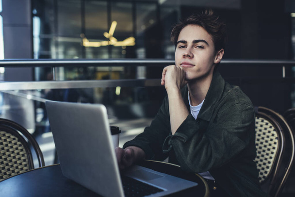 Young pensive male remote worker in casual outfit with hand at chin looking at camera while browsing internet on laptop - Foto, Imagem