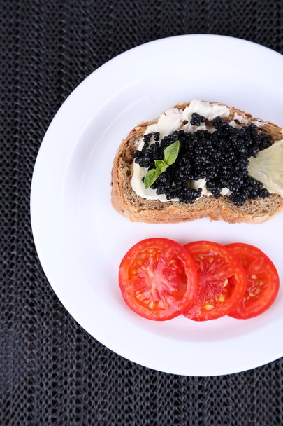 Black caviar and butter on bread with tomato on plate on dark fabric background - Фото, изображение