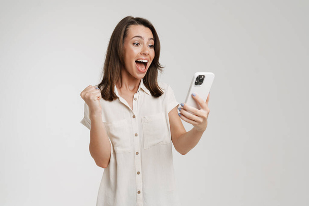 Young woman making winner gesture and using cellphone isolated over white background - Zdjęcie, obraz