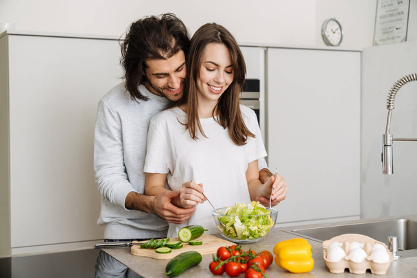 Young multiracial couple hugging while making salad together at home - Photo, Image