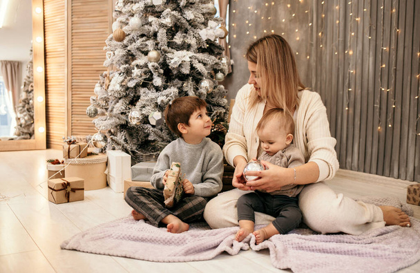 Blonde Mutter mit kleinen Söhnen sitzt auf einer Decke neben dem Weihnachtsbaum im Zimmer - Foto, Bild