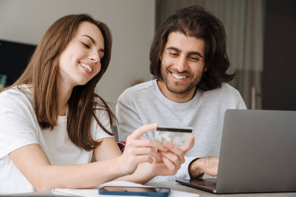 Young multiracial couple using credit card while working with laptop together at home - Photo, Image