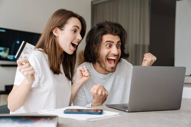 Young multiracial couple making winner gesture while using laptop together at home - Photo, Image