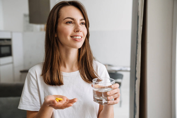 Smiling young brunette woman holding pills and glass of water whilastanding at home - Foto, afbeelding