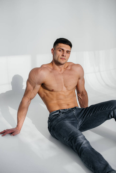 A young sexy athlete with perfect abs sits on the floor in the studio topless in jeans in the background. Healthy lifestyle, proper nutrition, training programs and nutrition for weight loss. - Fotografie, Obrázek