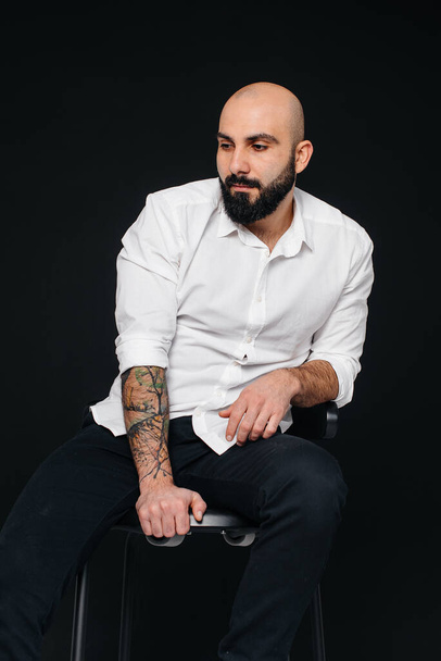 A young bearded man in a white shirt sits on a black background. - Foto, immagini