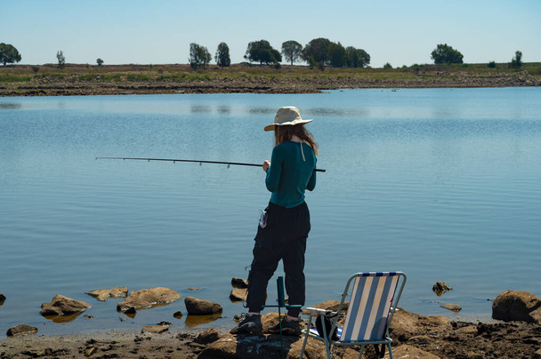 Ένα κορίτσι με ψάθινο καπέλο με καλάμι ψαρέματος στο χέρι στέκεται στην ακτή και ποζάρει μια ηλιόλουστη μέρα - Φωτογραφία, εικόνα