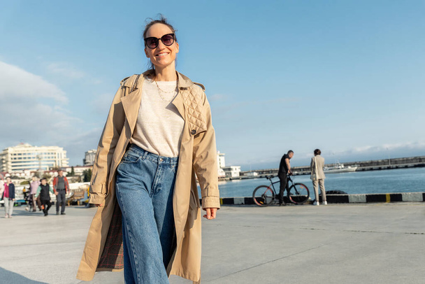 Молоді дорослі модні стилістичні красиві кавказькі щасливі усміхнені жінки люблять гуляти в Ялтинському морі в теплий сонячний день. Портрет жінки носить джинси бігове пальто на міській вулиці - Фото, зображення