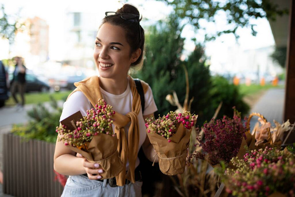 Photo of positive happy caucasian young brunette woman chooses potted beautiful flowers to buy at outdoor garden stall in the city - Фото, зображення