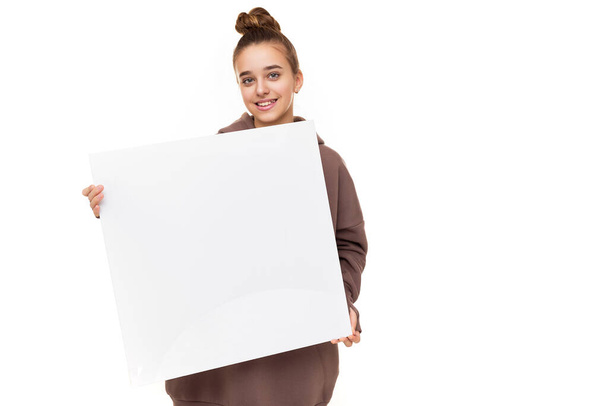 Photo of a beautiful positive smiling cute girl with dark hair and a bun in a brown hoodie with hood holding a large white square sheet with blank space isolated on white background with free space - Zdjęcie, obraz