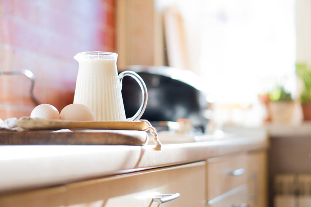 Φρέσκο γάλα και αυγά σε ξύλινη σανίδα σε πραγματικό σπίτι κουζίνα εσωτερικό με φυσικό φως - Φωτογραφία, εικόνα