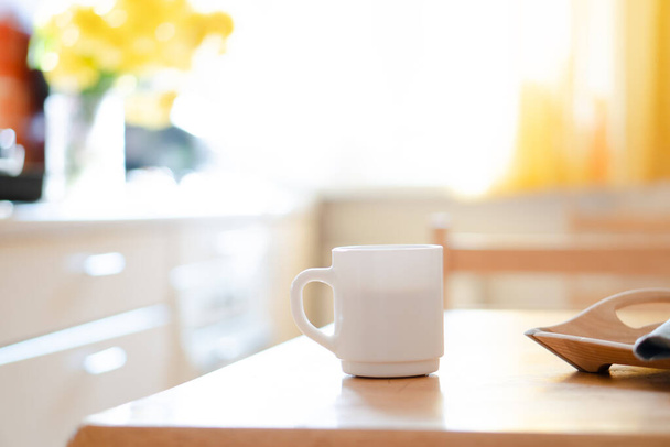 Bílý hrnek na kávu s ubrouskem na stole v reálném kuchyňském interiéru s jasným přírodním slunečním světlem - Fotografie, Obrázek